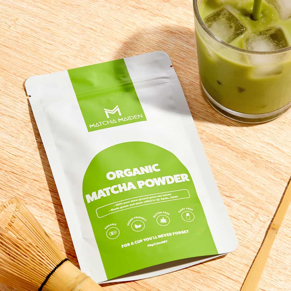 Matcha Maiden bamboo scoop and bamboo whisk on a wooden table with pouch of organic matcha powder and a refreshing iced.