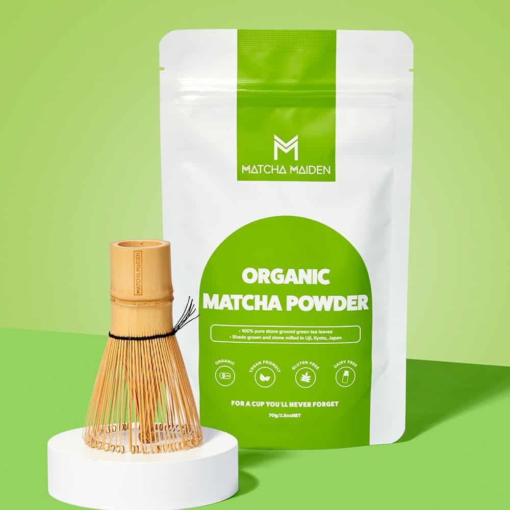 Matcha Maiden bamboo whisk standing upright on a white podium by 70g pouch of Organic Matcha Powder.