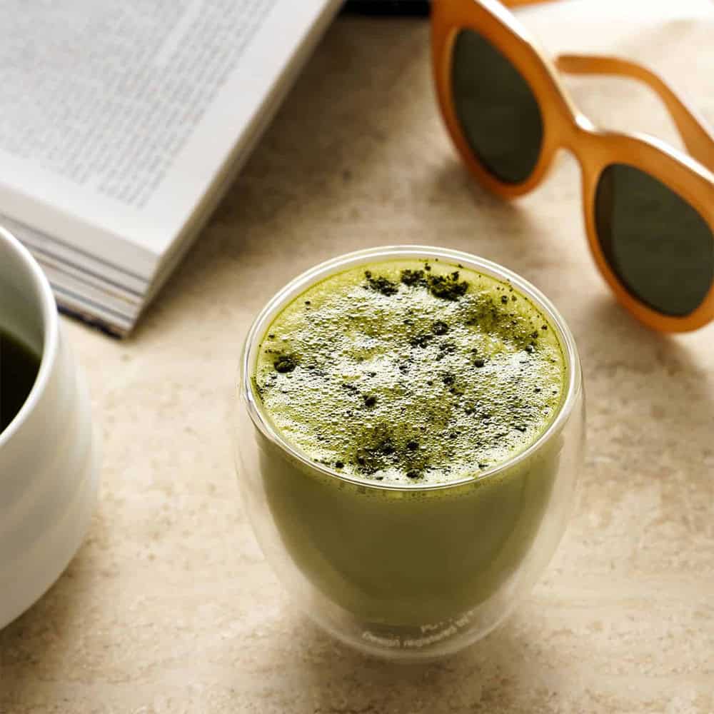 A frothy hot matcha latte dusted with green powder in a double-walled glass cup, styled with a book and orange sunglasses.