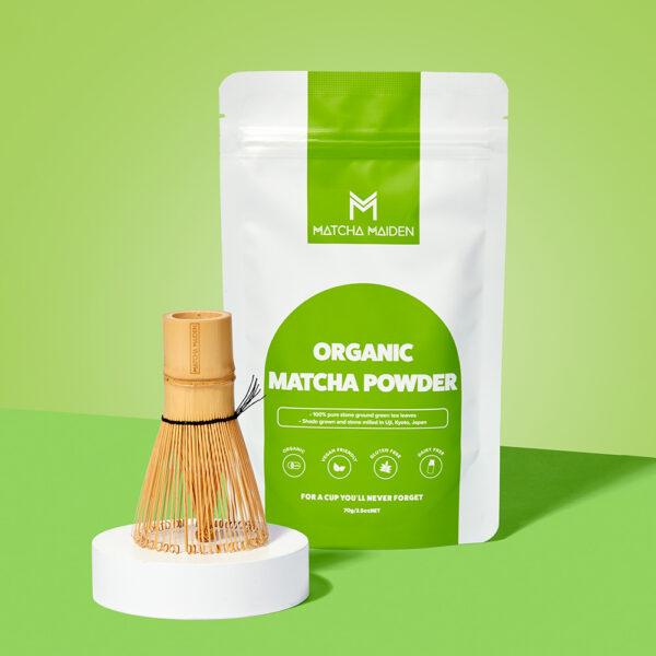 Matcha Maiden 70g Organic Matcha pouch displayed with traditional bamboo whisk on a white podium against.