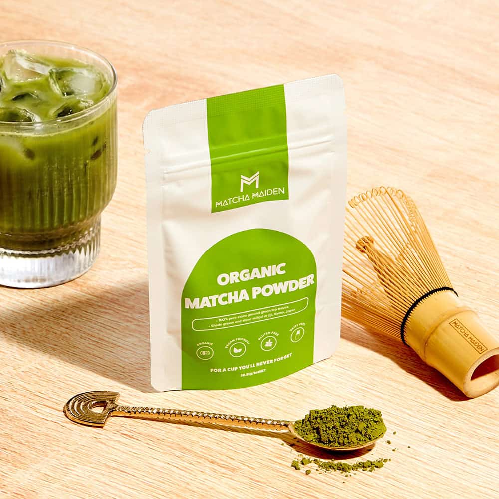 Matcha Maiden 28.35g organic matcha pouch on light wooden table with refreshing iced matcha latte and a bamboo whisk.