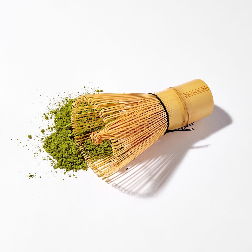 Matcha Maiden traditional bamboo whisk, also known as a chasen, on pile of vibrant green organic matcha powder.