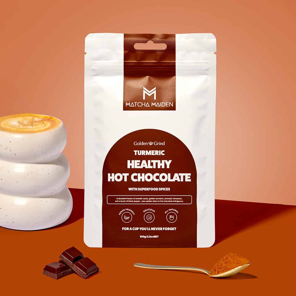 Matcha Maiden Golden Grind Turmeric Healthy Hot Chocolate 100g pouch by warm ribbed mug, dark chocolate squares.