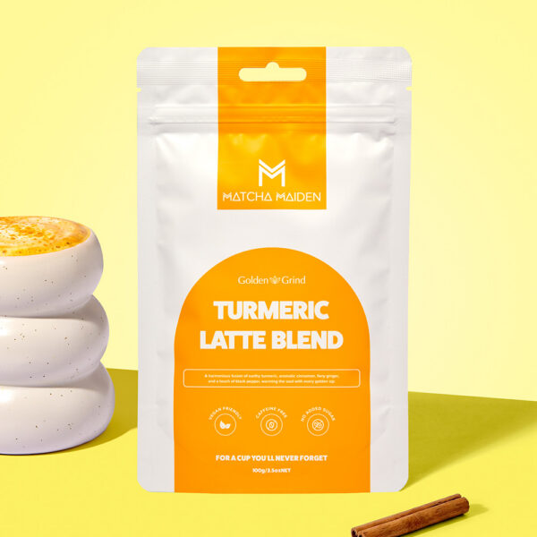 Matcha Maiden Golden Grind Turmeric Latte Blend 100g pouch by white ribbed mug filled with golden latte and a cinnamon stick.