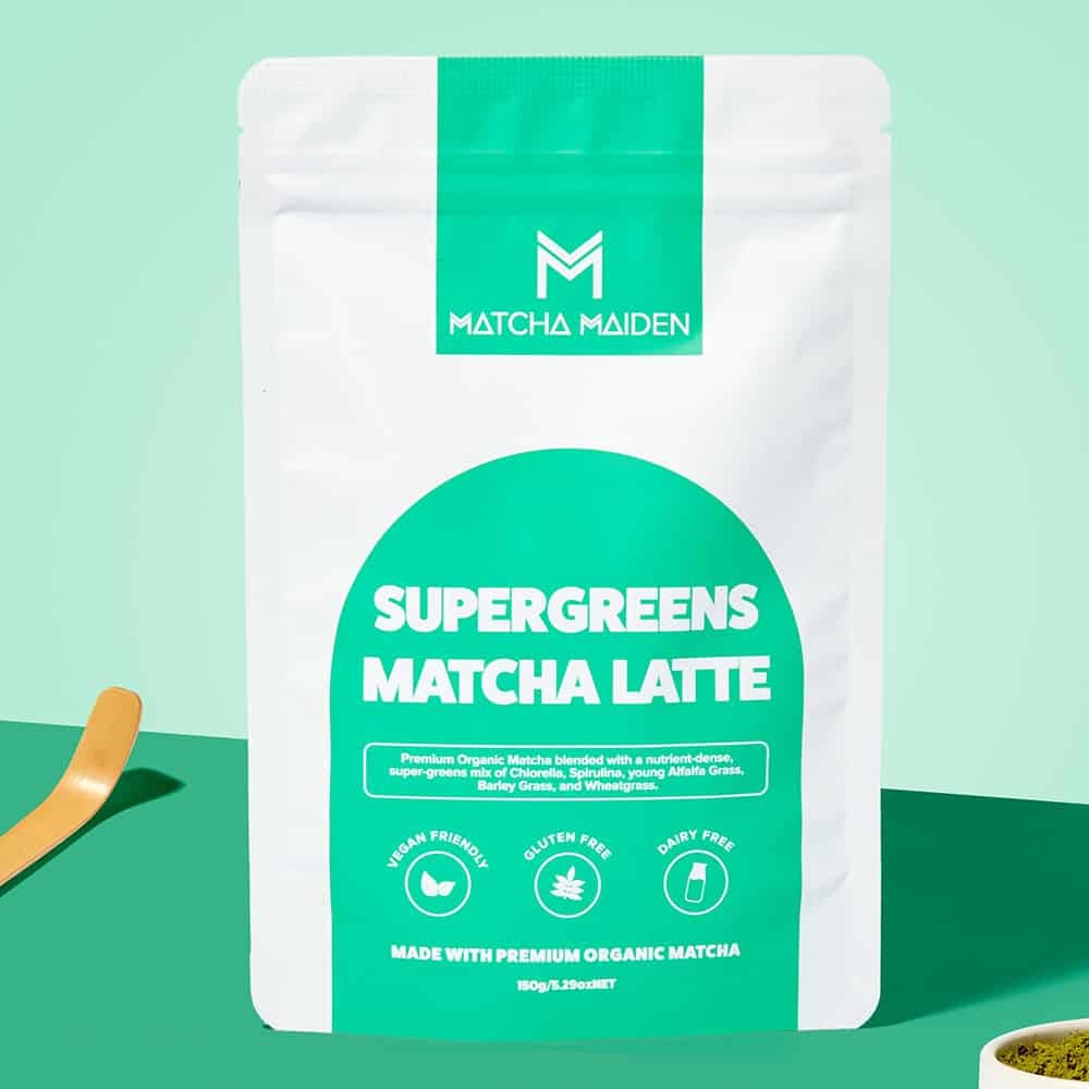 Matcha Maiden Supergreens Matcha Latte 150g pouch by a bamboo scoop and a bowl of green powder on a vibrant teal background.